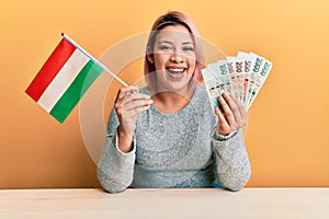 Hispanic woman with pink hair holding czech republic flag and koruna banknotes smiling and laughing hard out loud because funny