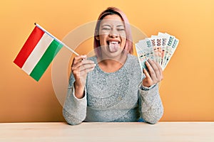 Hispanic woman with pink hair holding czech republic flag and koruna banknotes sticking tongue out happy with funny expression