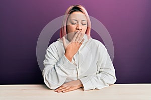 Hispanic woman with pink hair wearing casual sweatshirt sitting on the table bored yawning tired covering mouth with hand