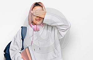 Hispanic woman with pink hair wearing student backpack and headphones covering eyes with hand, looking serious and sad