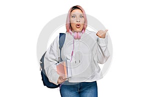 Hispanic woman with pink hair wearing student backpack and headphones surprised pointing with hand finger to the side, open mouth