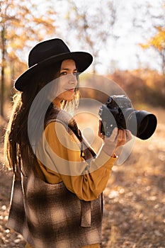 Hispanic woman is a professional photographer in black hat with dslr camera, outdoor in autumn forest,