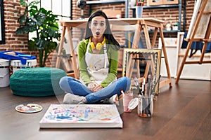 Hispanic woman sitting at art studio painting on canvas scared and amazed with open mouth for surprise, disbelief face