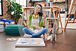 Hispanic woman sitting at art studio painting on canvas smiling happy pointing with hand and finger to the side