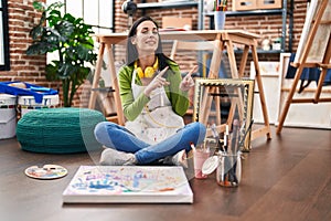 Hispanic woman sitting at art studio painting on canvas smiling and looking at the camera pointing with two hands and fingers to