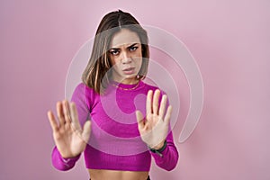 Hispanic woman standing over pink background moving away hands palms showing refusal and denial with afraid and disgusting