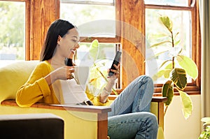 Hispanic woman using her smartphone and drinking coffee sitting in front of a window in a bright living room. Side