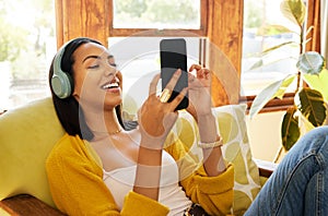 Hispanic woman using her smartphone and listening to music on headphones while comfortable and relaxing in a bright