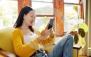 Hispanic woman using her smartphone sitting in front of a window in a bright living room. A young happy female smiling