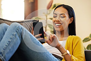 Hispanic woman using her smartphone while smiling and relaxing in a bright living room. A young female lying on a sofa