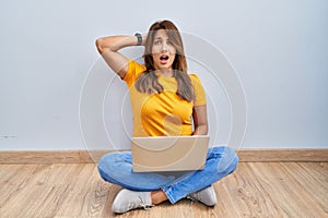 Hispanic woman using laptop sitting on the floor at home crazy and scared with hands on head, afraid and surprised of shock with