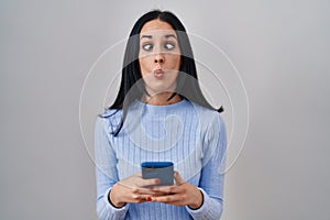 Hispanic woman using smartphone making fish face with mouth and squinting eyes, crazy and comical