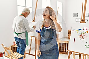 Hispanic woman wearing apron at art studio smiling with hand over ear listening an hearing to rumor or gossip