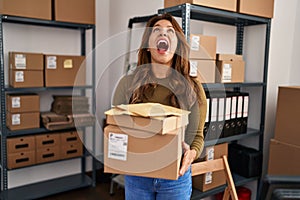 Hispanic woman working at small business ecommerce angry and mad screaming frustrated and furious, shouting with anger looking up