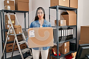 Hispanic woman working at small business ecommerce holding box skeptic and nervous, frowning upset because of problem