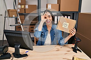 Hispanic woman working at small business ecommerce holding box smiling happy doing ok sign with hand on eye looking through