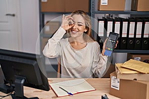 Hispanic woman working at small business ecommerce holding credit card and dataphone smiling happy doing ok sign with hand on eye
