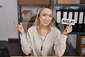 Hispanic woman working at small business ecommerce holding help banner smiling happy pointing with hand and finger to the side