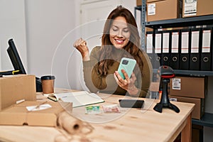 Hispanic woman working at small business using smartphone screaming proud, celebrating victory and success very excited with