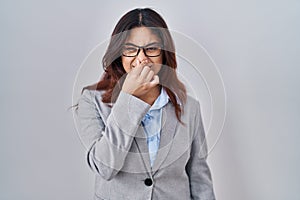 Hispanic young business woman wearing glasses smelling something stinky and disgusting, intolerable smell, holding breath with