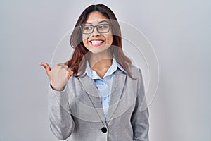 Hispanic young business woman wearing glasses smiling with happy face looking and pointing to the side with thumb up