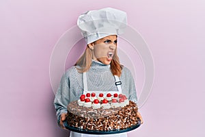 Hispanic young chef woman holding chocolate cake angry and mad screaming frustrated and furious, shouting with anger