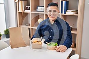 Hispanic young man eating take away food looking positive and happy standing and smiling with a confident smile showing teeth