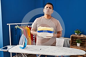 Hispanic young man holding folded laundry after ironing looking at the camera blowing a kiss being lovely and sexy