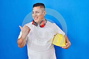 Hispanic young man holding hardhat pointing thumb up to the side smiling happy with open mouth