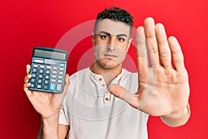 Hispanic young man showing calculator device with open hand doing stop sign with serious and confident expression, defense gesture