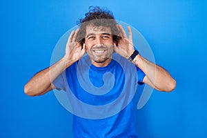 Hispanic young man standing over blue background trying to hear both hands on ear gesture, curious for gossip