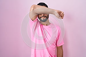 Hispanic young man standing over pink background covering eyes with arm, looking serious and sad