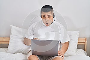 Hispanic young man using computer laptop on the bed scared and amazed with open mouth for surprise, disbelief face