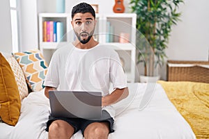 Hispanic young man using computer laptop sitting on the bed scared and amazed with open mouth for surprise, disbelief face