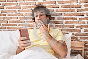 Hispanic young man using smartphone on the bed covering mouth with hand, shocked and afraid for mistake