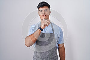 Hispanic young man wearing apron over white background asking to be quiet with finger on lips