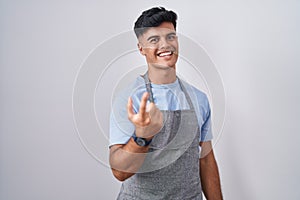 Hispanic young man wearing apron over white background beckoning come here gesture with hand inviting welcoming happy and smiling
