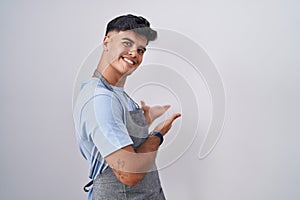 Hispanic young man wearing apron over white background inviting to enter smiling natural with open hand
