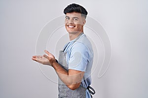 Hispanic young man wearing apron over white background pointing aside with hands open palms showing copy space, presenting