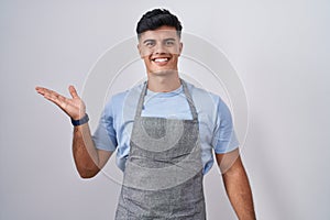 Hispanic young man wearing apron over white background smiling cheerful presenting and pointing with palm of hand looking at the