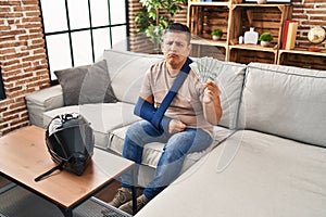 Hispanic young man wearing arm on sling holding usa dollars banknotes depressed and worry for distress, crying angry and afraid