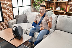 Hispanic young man wearing arm on sling holding usa dollars banknotes smiling and laughing hard out loud because funny crazy joke