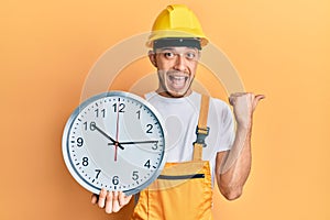 Hispanic young man wearing builder uniform and hardhat holding clock pointing thumb up to the side smiling happy with open mouth