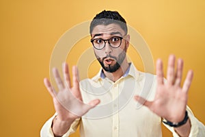 Hispanic young man wearing business clothes and glasses afraid and terrified with fear expression stop gesture with hands,