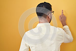 Hispanic young man wearing business clothes and glasses posing backwards pointing ahead with finger hand