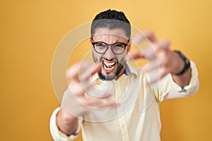 Hispanic young man wearing business clothes and glasses shouting frustrated with rage, hands trying to strangle, yelling mad