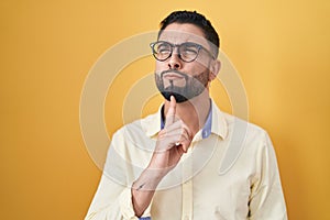 Hispanic young man wearing business clothes and glasses thinking concentrated about doubt with finger on chin and looking up