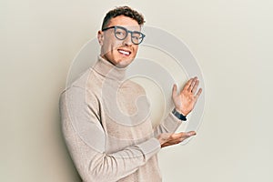 Hispanic young man wearing casual turtleneck sweater inviting to enter smiling natural with open hand