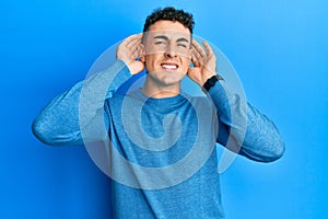 Hispanic young man wearing casual winter sweater trying to hear both hands on ear gesture, curious for gossip