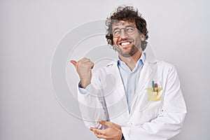 Hispanic young man wearing doctor uniform smiling with happy face looking and pointing to the side with thumb up